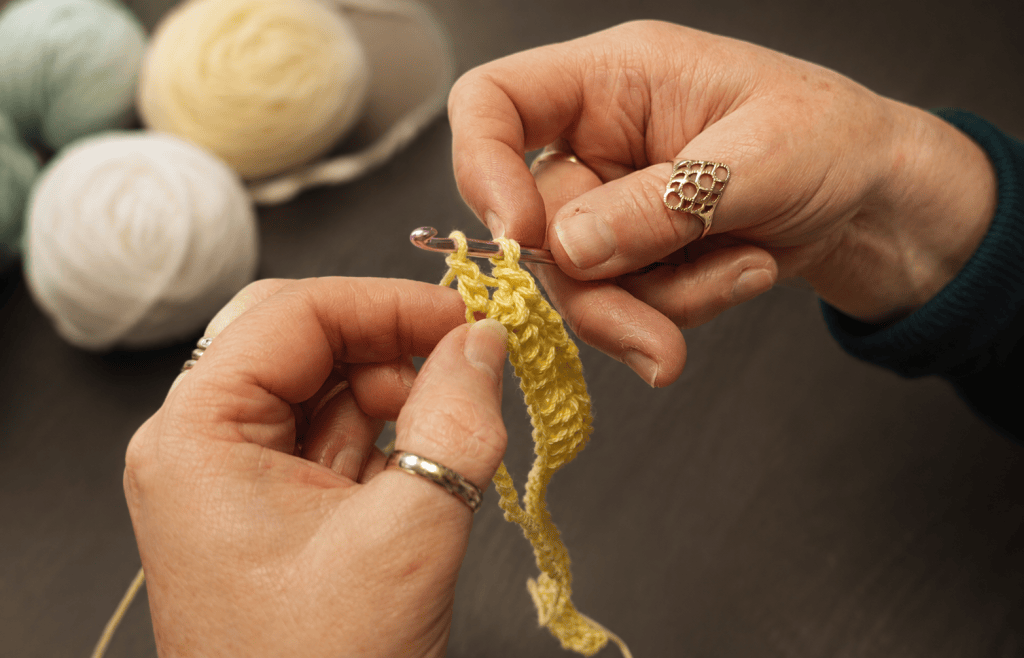 Hands crocheting a yellow piece with balls of yarn in the background.